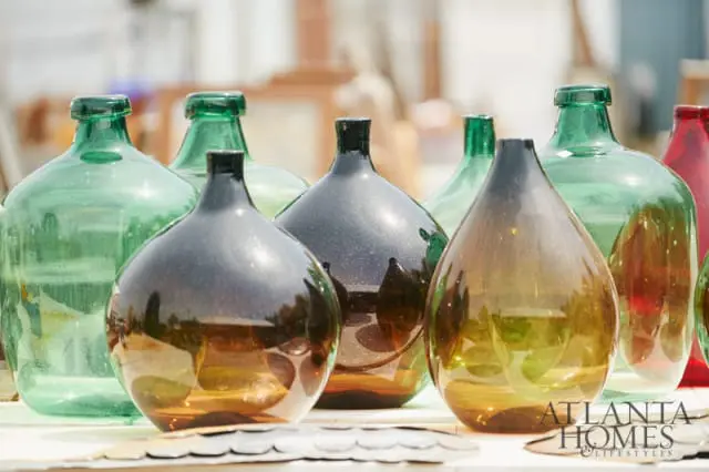 Collection of large glass demijohns in green, amber, and dark gray hues, with the Atlanta Homes & Lifestyles logo visible in the lower right corner.