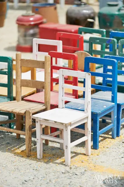 Row of colorful wooden chairs, including white, blue, red, and green, with weathered finishes, arranged outdoors on a gravel surface.