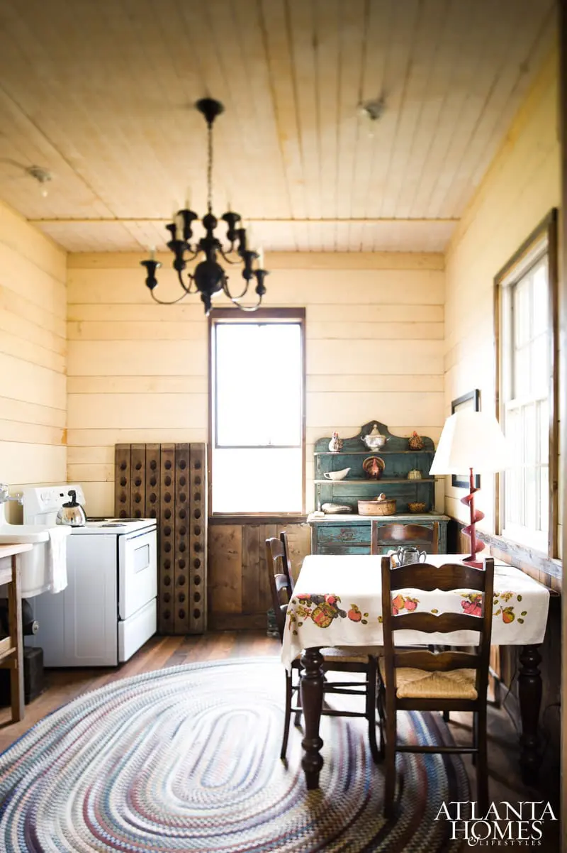 Rustic kitchen with wood-paneled walls and ceiling, white stove, oval braided rug, and dining table with floral tablecloth.