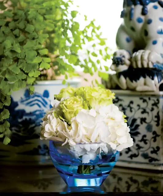 Close-up of a bouquet of white hydrangeas and pale green roses in a blue glass vase, with greenery and blue and white ceramic decor in the background.