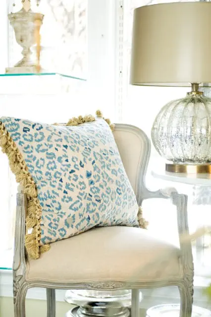 Cream upholstered armchair with a blue and white leopard print pillow with tan tassels. A mercury glass lamp sits on a table to the right.