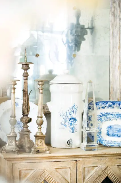 White ceramic water dispenser with blue Chinoiserie pattern, brass candlesticks, and blue and white plates on a distressed wood cabinet.