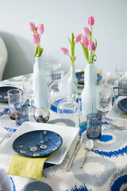 Table set with blue and white patterned tablecloth, white square plates, blue floral accent plates, yellow napkins, and pink tulips in light blue vases.