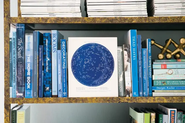 Bookshelf with a collection of books in shades of blue and white, a framed constellation chart, and gold decorative objects. Titles include 'The South,' 'The Home Book,' 'Rooms,' 'Decorating,' 'Thad Hayes,' and 'The Southern Cottage.'