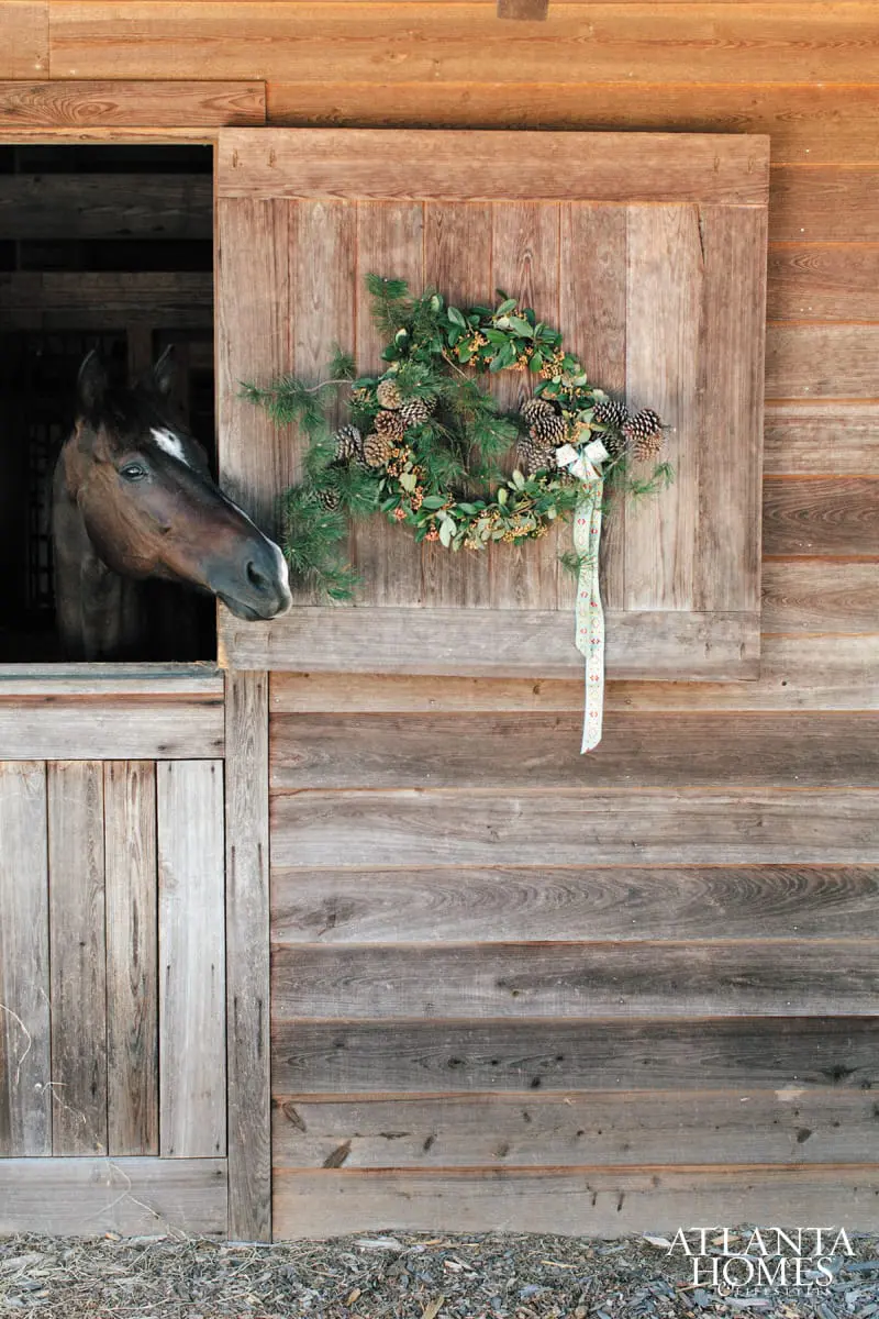 Brown horse peeking out of a barn stall next to a rustic wooden door adorned with a pinecone and greenery wreath.