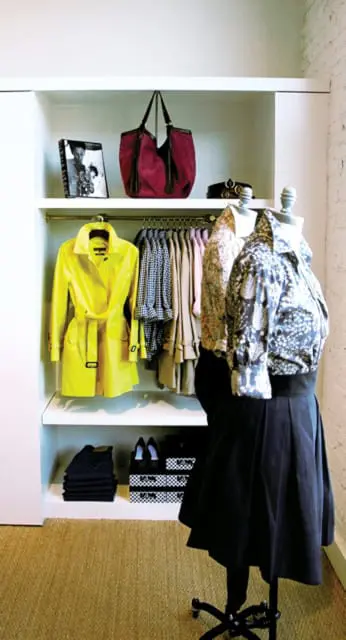 White shelving unit displaying a bright yellow trench coat, patterned shirts on mannequins, handbags, and folded dark denim jeans.