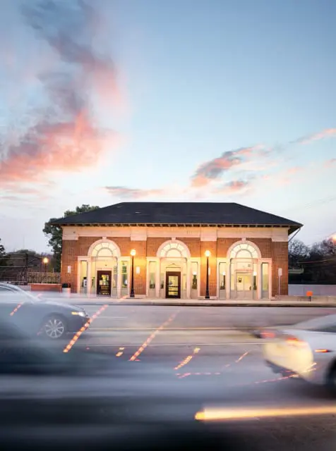 Brick train station with arched windows and doors, illuminated by streetlights at dusk, with blurred cars in the foreground.