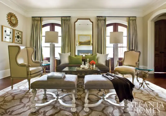 Living room with a dark brown sofa, green and white pillows, two floor lamps, two upholstered ottomans, and a large patterned rug.