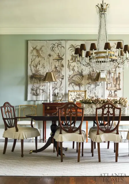 Dining room with a dark wood pedestal table and chairs featuring upholstered seats and carved backs. A large crystal chandelier hangs above. The wall is adorned with a triptych chinoiserie mural.