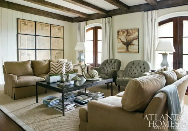 Living room with neutral-toned sofas, tufted armchairs, a dark wood coffee table with books, and a grid of framed maps on the wall. Exposed wooden beams accent the ceiling.