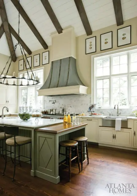 Cream kitchen with a large gray range hood, marble countertops, and a green island with a wooden breakfast bar. Exposed wooden beams accent the vaulted ceiling.