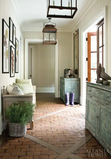 Hallway with brick floor, paneled walls, and rustic wooden furniture. Features include a bench with pillows, a distressed blue cabinet with bird figurines, and two hanging lanterns.