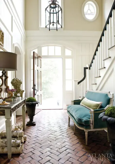 Entryway with herringbone brick floor, teal settee with green pillow, console table with bust and logs, and a large lantern hanging from the ceiling.