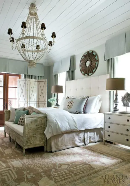 Bedroom with white tufted headboard, light blue accent pillows, distressed wood bench, ornate chandelier, and patterned rug.