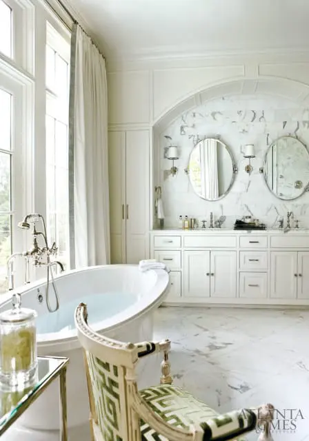 White bathroom with freestanding soaking tub, marble tile walls, double vanity with oval mirrors, and a decorative chair with green geometric upholstery.