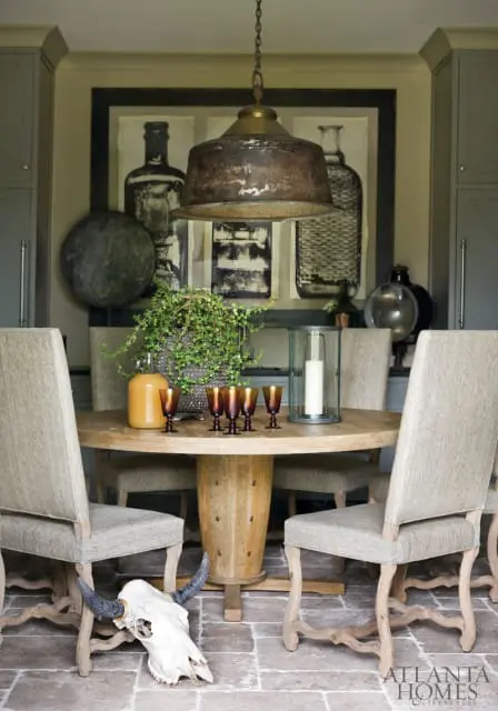 Round wooden dining table with four upholstered chairs, a large metal pendant light, and a framed artwork of bottles on the wall.