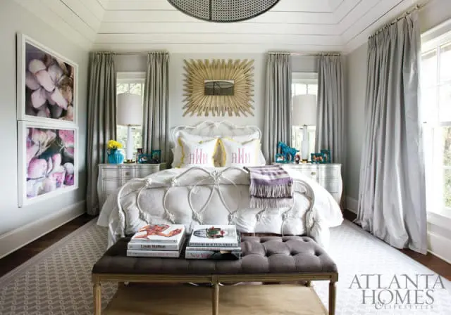 Bedroom with a white metal bed, monogrammed pillows, and a sunburst mirror above the headboard. Gray curtains frame the windows, and two framed floral prints hang on the wall.