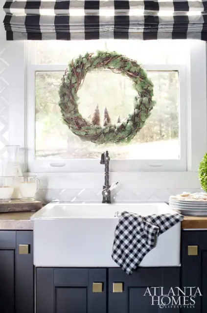 Farmhouse sink in a kitchen with dark blue cabinets, brass hardware, and a black and white buffalo check roman shade above the window. A moss wreath with miniature trees hangs in the window.
