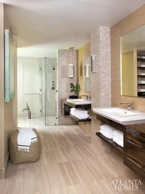 Modern bathroom with dual vanities featuring dark wood cabinets, white sinks, and stacked white towels. A glass-enclosed shower is visible in the background, with mosaic tile accents on the walls.