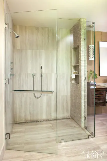 Modern bathroom with a large glass-enclosed shower featuring travertine tile walls and floor, a built-in bench, and dual shower heads. A vanity with dark wood cabinetry is visible to the right.