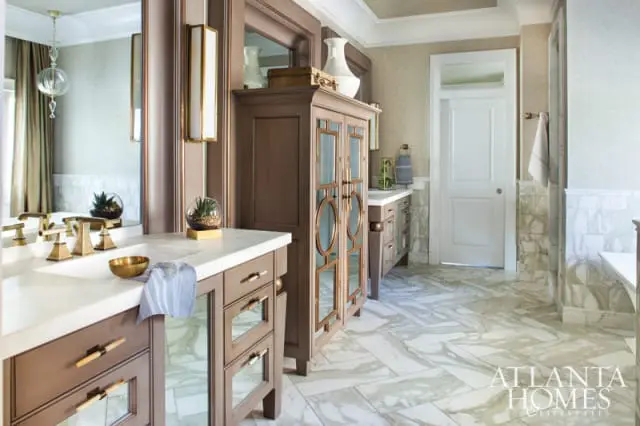 Luxury bathroom with marble floors, a double vanity with brass fixtures, a tall brown cabinet with mirrored doors, and a white door with a glass transom.