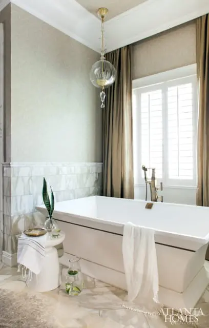 White freestanding bathtub with brass fixtures, marble tile wainscoting, textured wallpaper, and a glass globe chandelier.