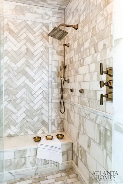 Marble tiled shower with herringbone pattern on the back wall, a built-in marble bench, and brushed bronze fixtures. Atlanta Homes & Lifestyles logo in the bottom right corner.