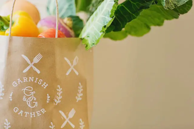 Brown paper grocery bag with white "Garnish & Gather" logo and cutlery icons, filled with fresh produce including yellow bell pepper, turnips, and leafy greens.