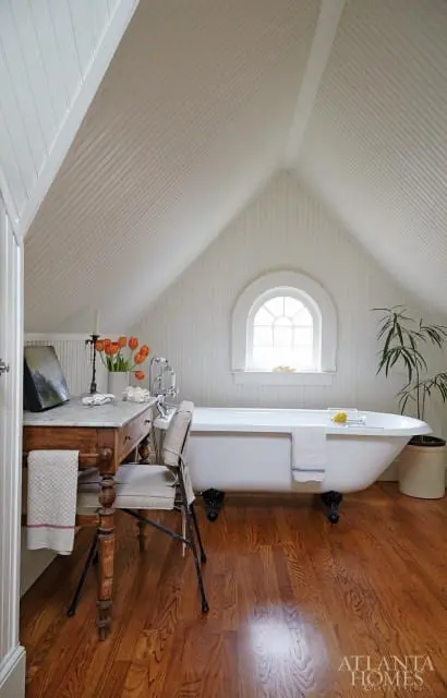 Attic bathroom with white beadboard walls and ceiling, clawfoot bathtub, arched window, and wooden vanity with marble top.