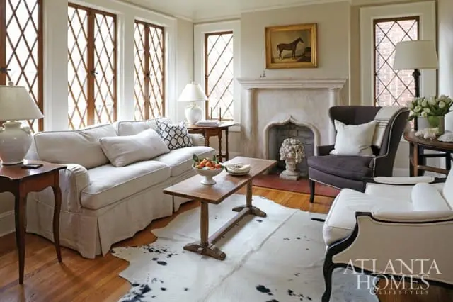 Living room with a white slipcovered sofa, dark upholstered armchair, and a white armchair. Features diamond-paned windows, a stone fireplace with a painting above, and a cowhide rug.