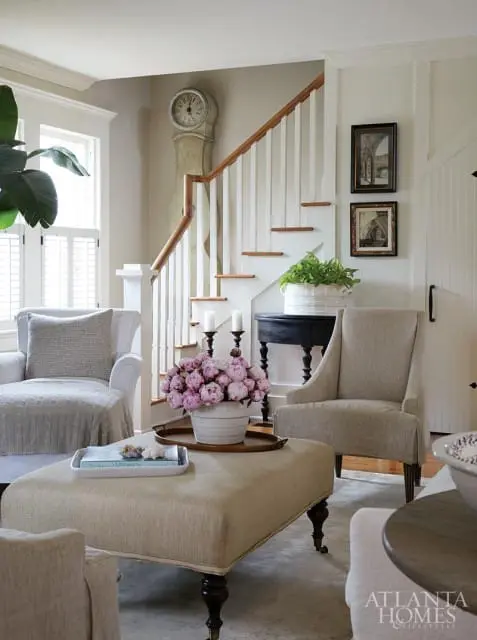 Living room with a light gray armchair, upholstered ottoman, and a beige armchair next to a staircase with a grandfather clock and framed artwork.