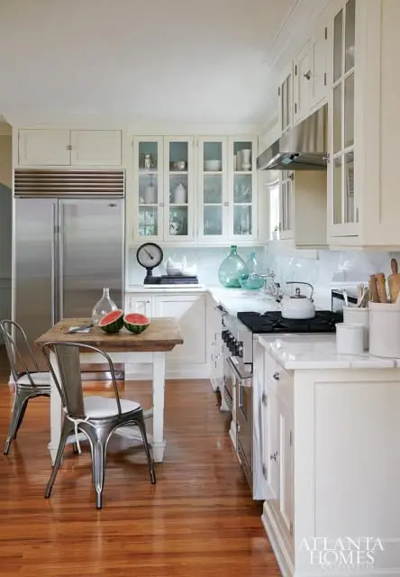 Cream kitchen with stainless steel refrigerator, glass-front cabinets, butcher block table with metal chairs, and a professional range.