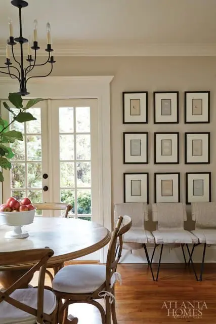 Dining area with a round wooden table, a white bowl of red apples, French doors leading outside, a black chandelier, and a wall of nine framed artworks above a bench with three upholstered seats.