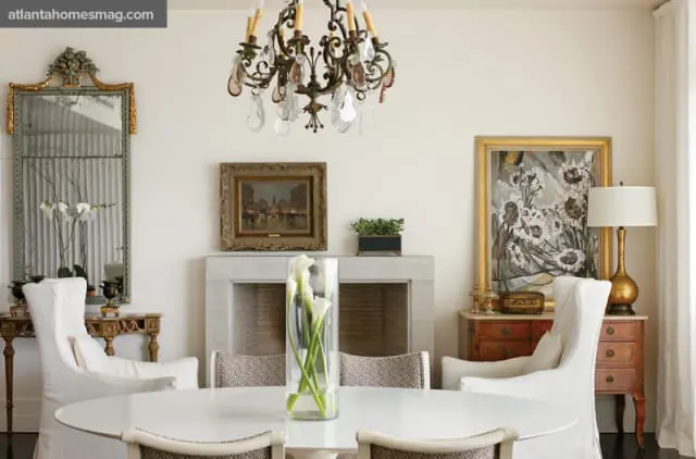 Dining room with a white oval table, white slipcovered chairs, a marble fireplace, and ornate mirrors and artwork.