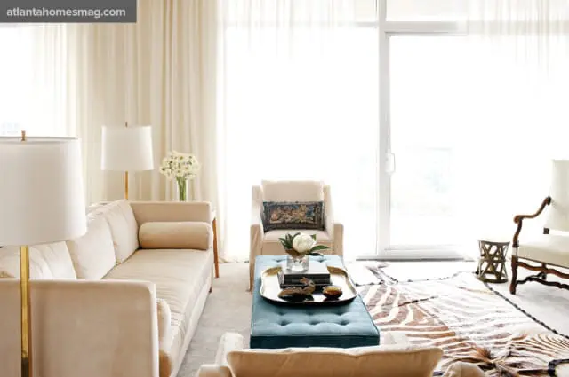 Living room with cream-colored velvet sofas, a teal tufted ottoman, a zebra print rug, and sheer floor-to-ceiling curtains.