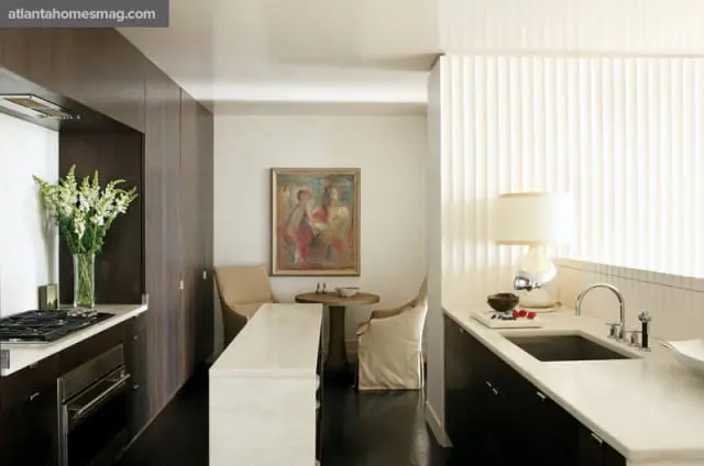 Modern kitchen with dark wood cabinetry, white marble countertops, and a central island. A painting hangs above a small dining table with two chairs.