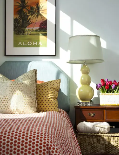 Bedroom with light blue headboard, patterned pillows, and a red and white polka dot duvet. A yellow lamp sits on a wooden nightstand next to a vase of tulips and a basket of yarn.
