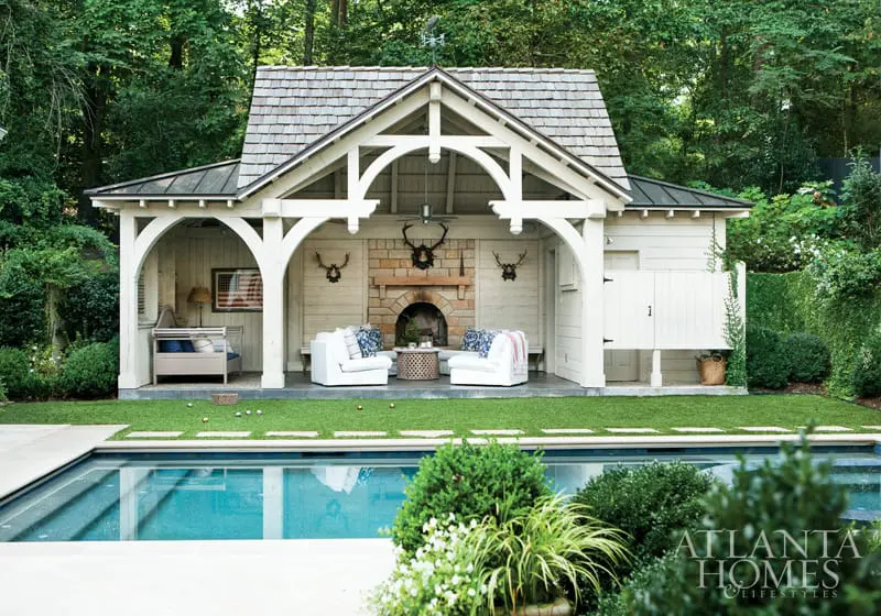 White pool house with stone fireplace, seating area, and adjacent swimming pool surrounded by lush greenery.
