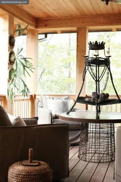 Covered porch with wooden ceiling and floors, featuring seating with neutral-toned upholstery, staghorn ferns mounted on a wooden post, and a metal table with a wire base holding decorative artichokes.