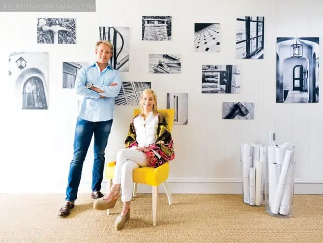 A man and woman stand and sit in front of a white paneled wall adorned with black and white photographs of architectural details.
