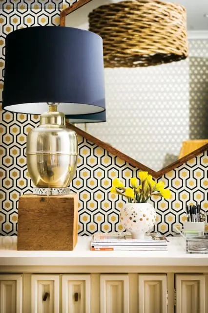 Gold metallic lamp with navy blue shade on a wooden block, next to a white vase with yellow flowers and magazines.
