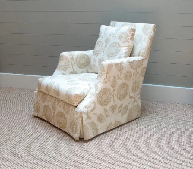 Armchair upholstered in cream fabric with a gold floral pattern, featuring a skirted base and two throw pillows. The chair sits on a textured beige carpet in front of a grey paneled wall.