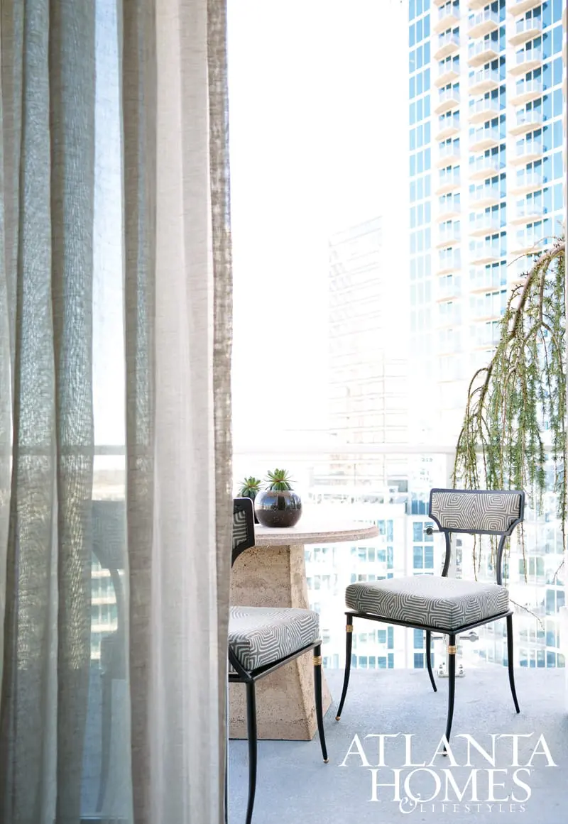 Balcony with two chairs featuring geometric patterned upholstery and a stone pedestal table with succulents, overlooking city skyscrapers.