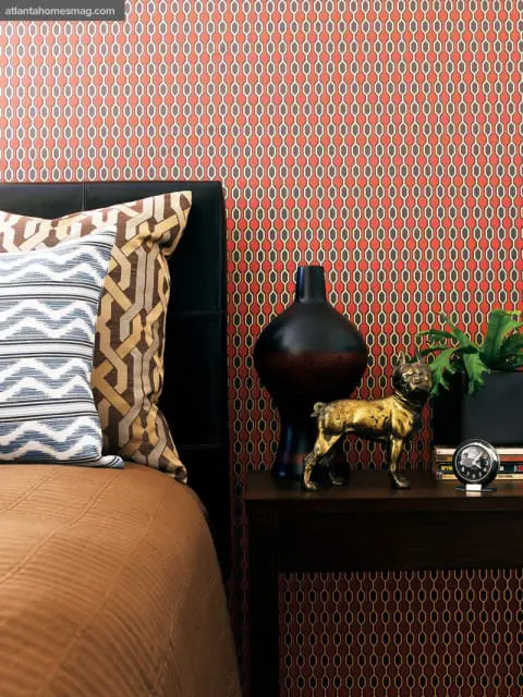 Bedroom with a black leather headboard, patterned wallpaper in red, grey, and white, and a bedside table with a black vase, brass dog figurine, and alarm clock.