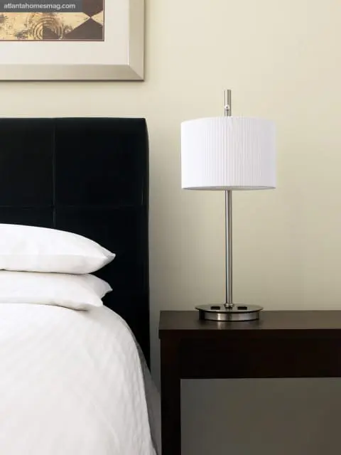 Bedroom with a black tufted headboard, crisp white bedding, and a modern silver lamp with a white pleated shade on a dark wood nightstand.