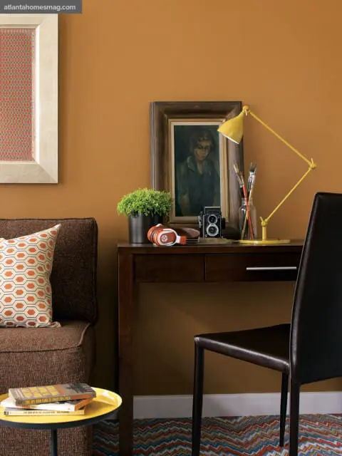 Brown sofa with geometric patterned pillow next to a dark wood desk with a yellow lamp, vintage camera, and framed painting.