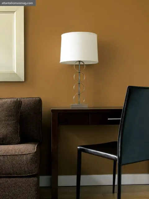 Desk with a modern lamp and a dark leather chair against a warm brown wall, with a textured brown sofa and a silver-framed mirror to the left.