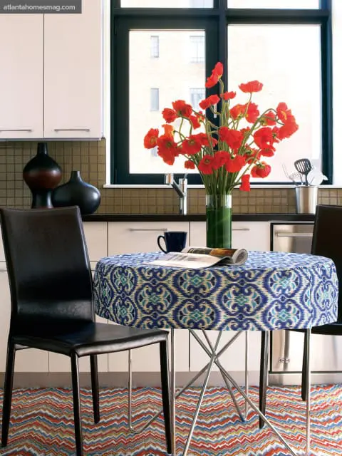 Modern kitchen with white cabinetry, mosaic tile backsplash, and a round table covered in a blue ikat print fabric. Red poppies in a vase sit on the counter.