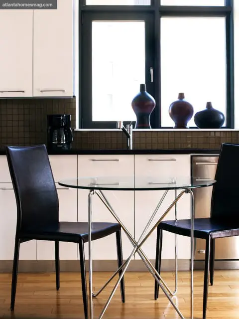Modern kitchen with cream cabinetry, dark tiled backsplash, and a glass-top dining table with two black leather chairs.