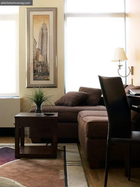 Living room with a brown sectional sofa, dark wood side table with a glass vase of grass, and a framed print of the Empire State Building on a cream wall.